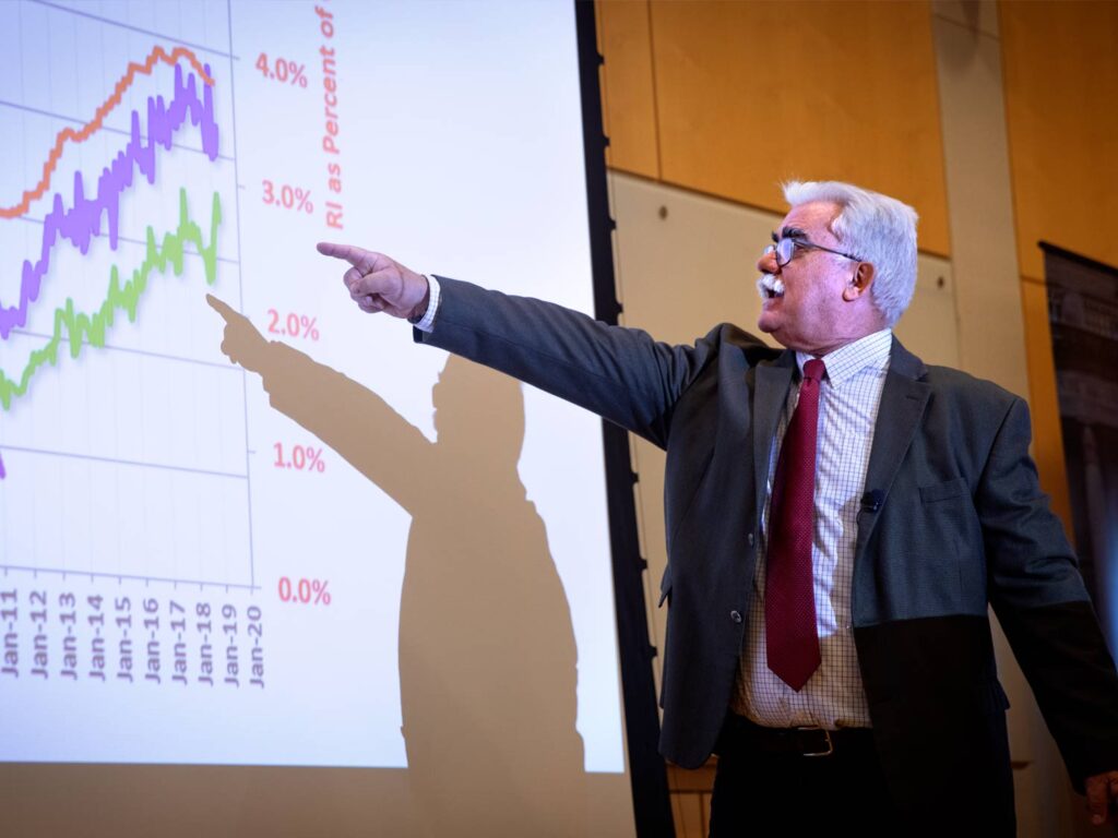 Male lecturer stands before a projector screen pointing to a chart on the screen.