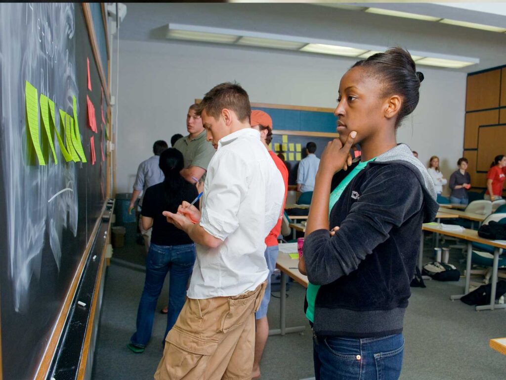 Students in a classroom write on sticky notes and place them on the chalkboard.