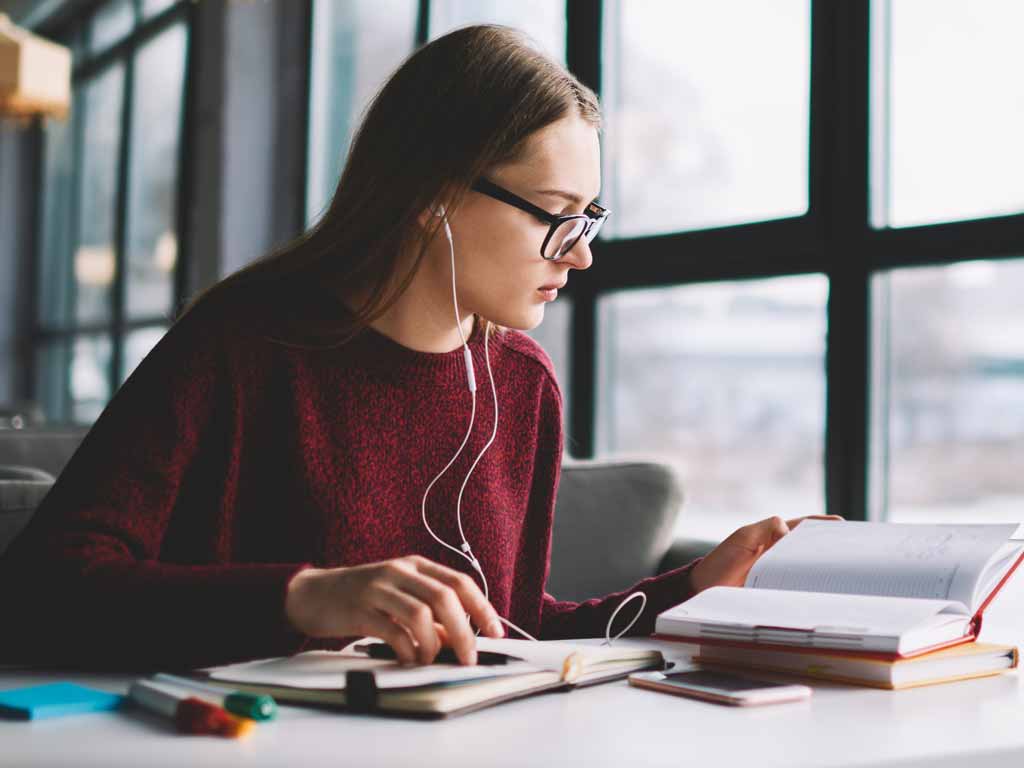 A woman wearing glasses sitting at a table with headphones in and three books open in front of her.