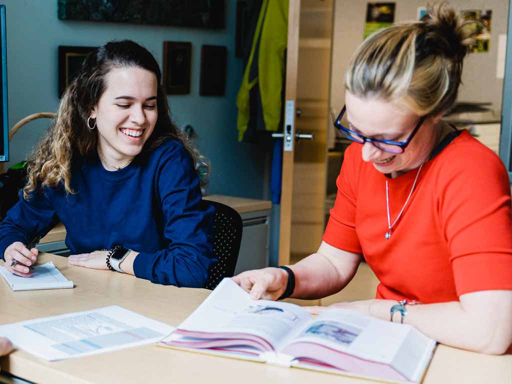 Two women sitting at a desk smiling while reading a textbook and taking notes.