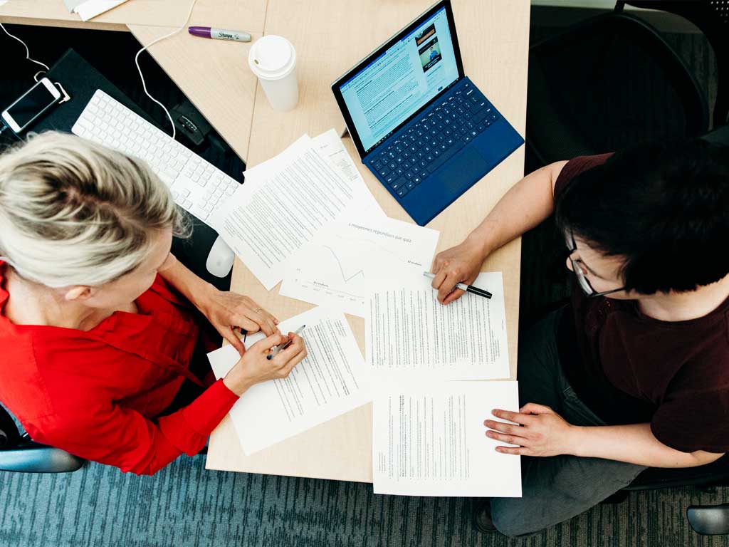 A professor and student sit across from each other at a desk reviewing printed papers.