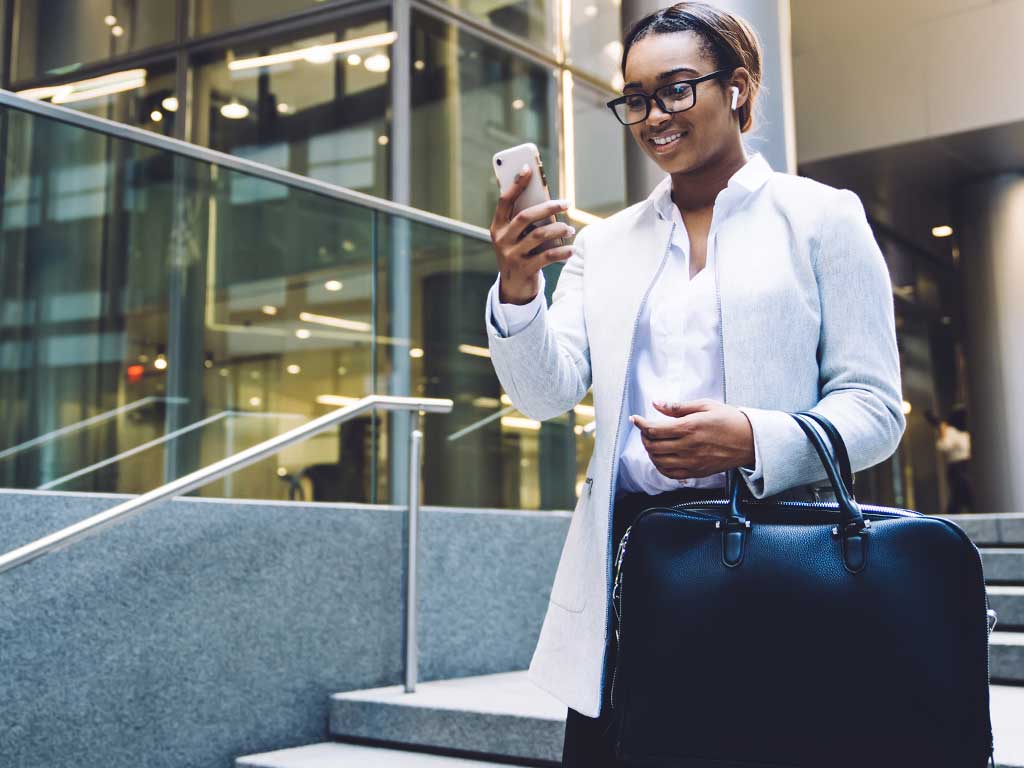 A woman in business attire holding a briefcase checks her phone in a metropolitan area.