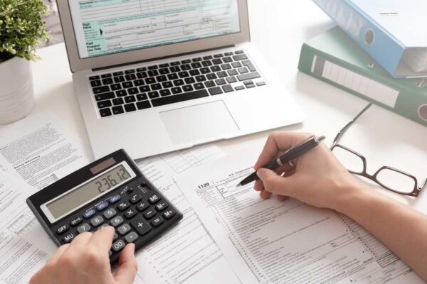 A person filling out tax forms with a calculator, laptop, and binders.