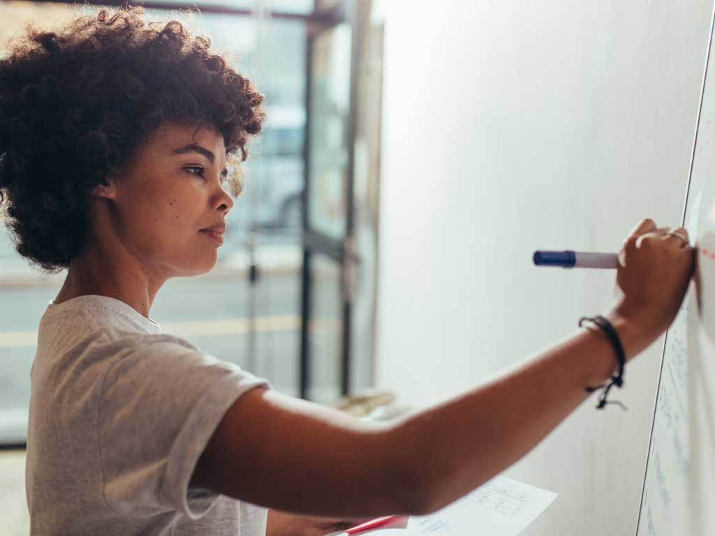 Woman in a white t-shirt writing on a whiteboard.