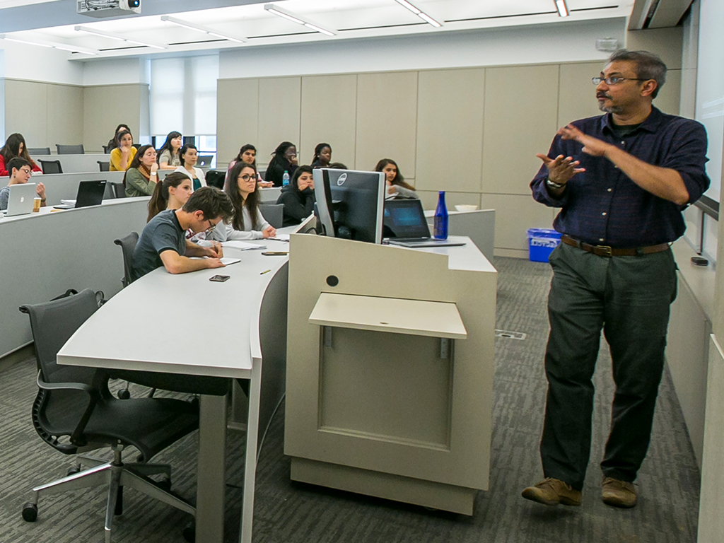 A professor stands in front of a tiered classroom teaching.