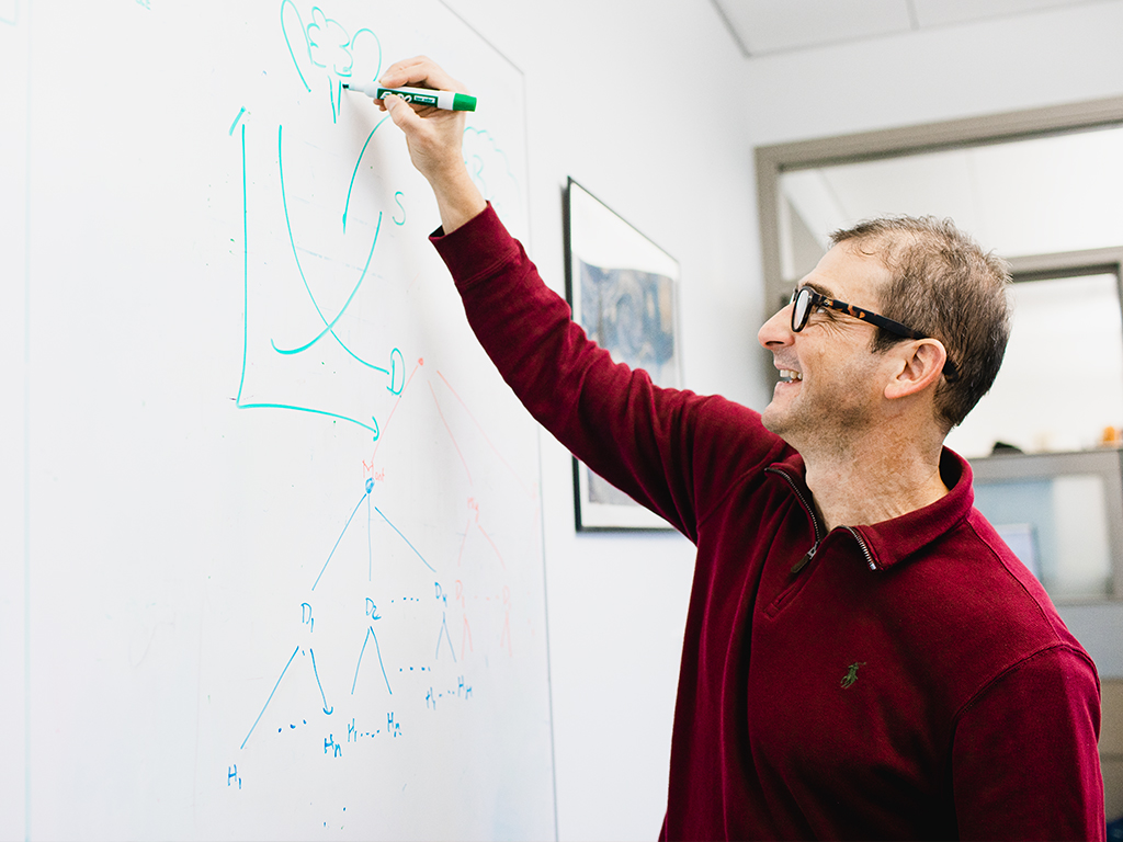 A professor wearing a red sweater writes on a whiteboard.
