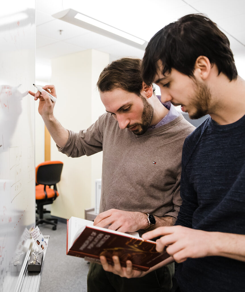 Two men stand at a whiteboard reading a textbook and writing on the board.