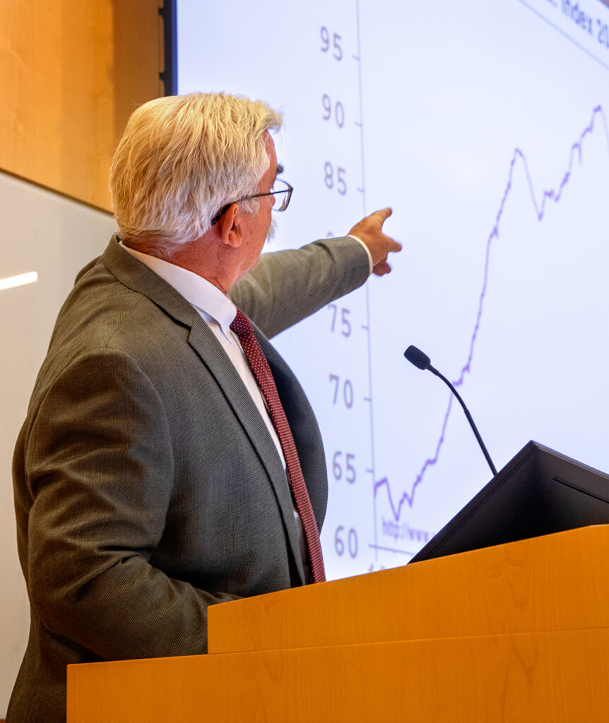 A man in a suit stands at a podium and points to a chart on a projector screen.