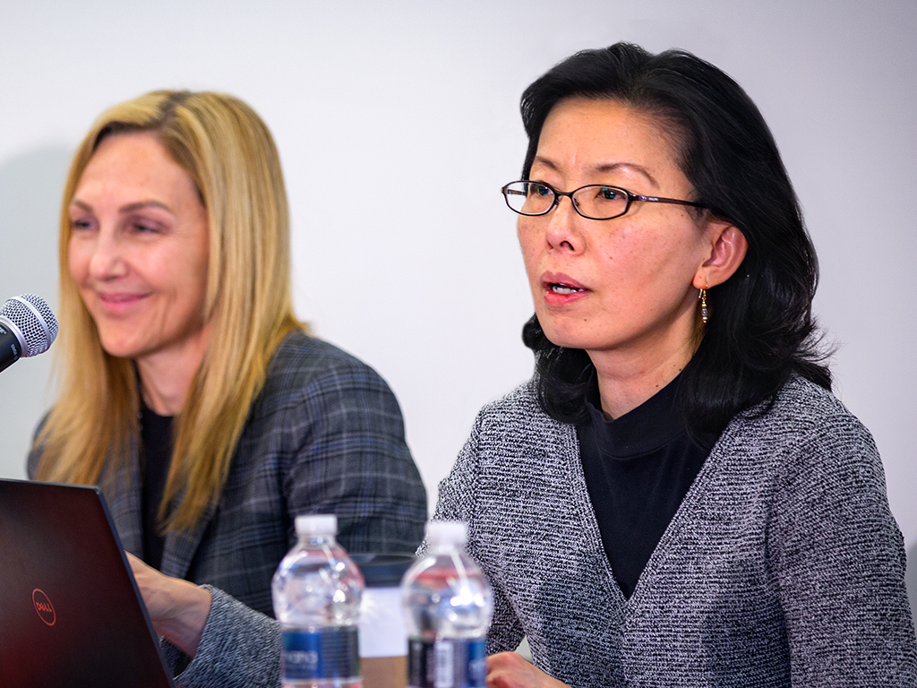 Two women sit at a table with a laptop and microphone in front of them