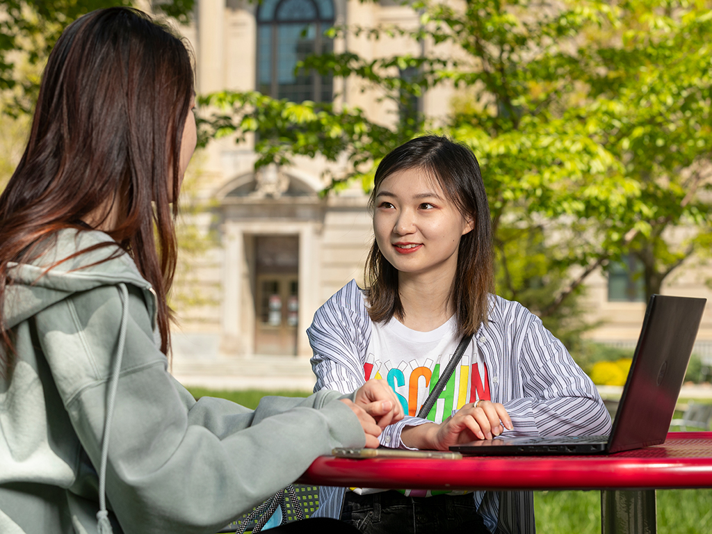 Two women sit outside at a picnic table with a laptop in front of them and Warren Hall visible in the background.
