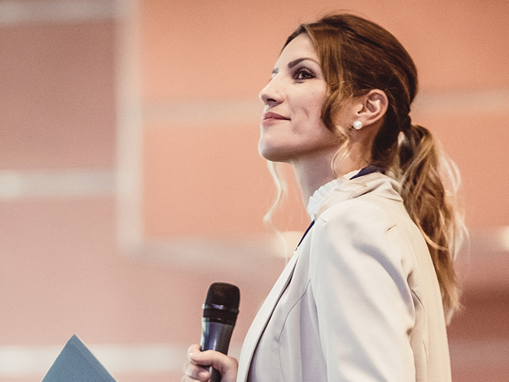 Profile of a woman in a blazer holds a microphone and looks to the side.